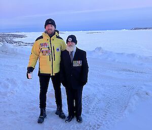 A man in yellow outdoor gear and a man wearing military medals stand in the snow