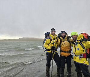 Three men in field gear, wearing field packs, stand on the side of a lake and smile to camera