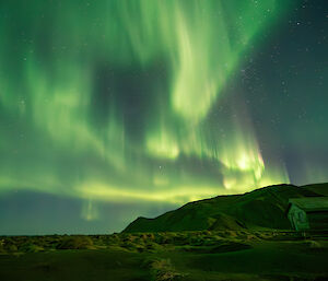 South end of station with unused hut and mountains behind with bright green aurora dancing in the sky above