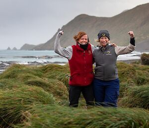 Two women in wind amongst tussocks raise arms in salute and smile at the camera