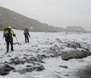 Two people with packs on walk away from camera across snow covered beach with heavy snow falling