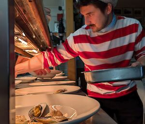 Man in stripy shirt and beret helps serve seafood onto bowls on a bench under a heat lamp