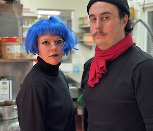 Two people dressed in black turtlenecks, one with blue hair, one with red scarf and black beanie, stare at the camera