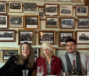 Three people sit at the table with a wall of framed photographs behind them
