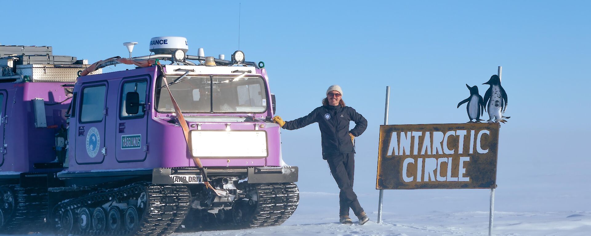 Alex stands next to the Antarctic circle sign with purple hagg, a snowy foreground with clear sky in the background