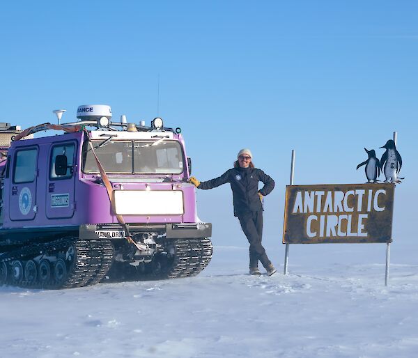 Alex stands next to the Antarctic circle sign with purple hagg, a snowy foreground with clear sky in the background