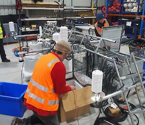 Three people working on a disassembled scientific tower lying horizontally on the floor of a warehouse