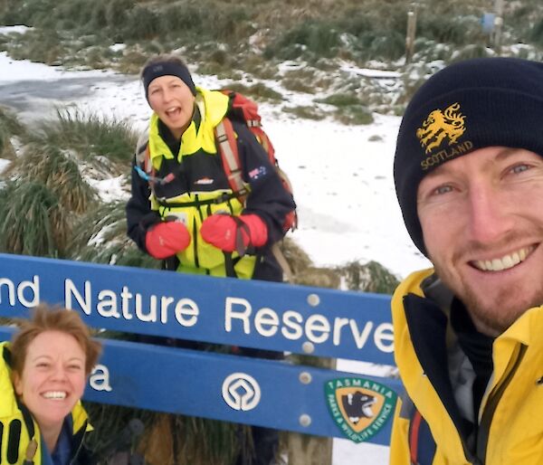 Three people stand around Macquarie island Nature Reserve Sign in field clothing and carrying field packs