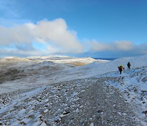 Two people in the distance walking across a snow covered hilly landscape with blue skies above