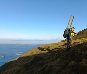 Woman wearing field pack, with two wooden stakes sticking out of the top, stands on the edge of steep grassy slope that drops down to the sea in the distance