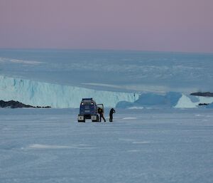 Beautiful winter blues and purples on a crisp cold morning off Mawson on the sea ice. Two expeditioners stand by a Hagglunds vehicle. One is drilling the sea-ice to determine thickness