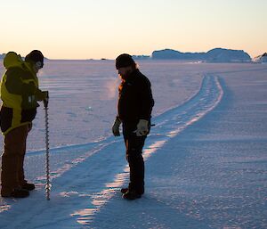 Two people stand on the ice with a long metal tool drilling into it.