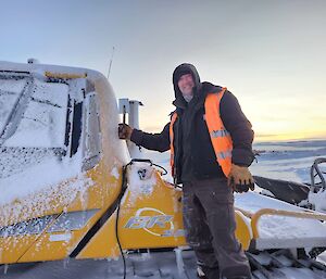 Expeditioner Dean Ahern is pictured with a large smile standing atop a snow groomer with a very bright sunrise in the background