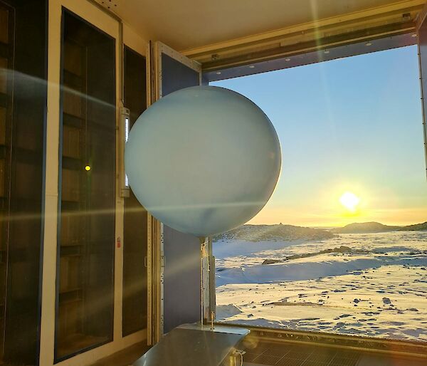 A weather balloon in a room in front of a window with a snowscape and blue sky outside