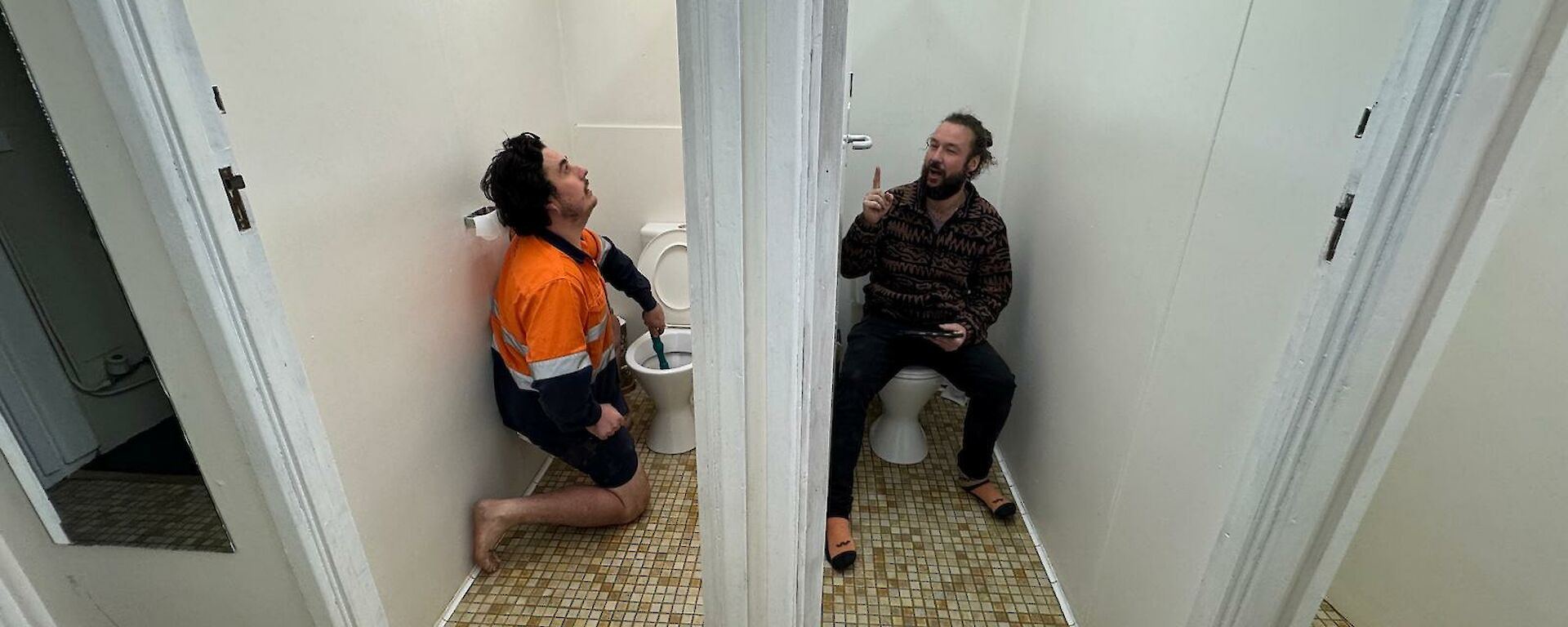 Two men in side by side toilet cubicles, on sitting on toilet and one working with plunger in toilet