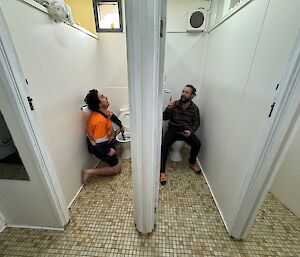 Two men in side by side toilet cubicles, on sitting on toilet and one working with plunger in toilet