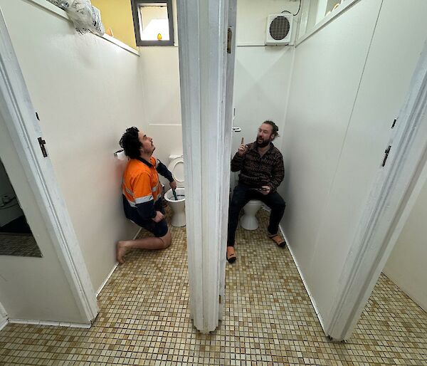 Two men in side by side toilet cubicles, on sitting on toilet and one working with plunger in toilet