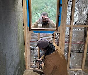 One man working on pipes in new building extension, with second looking on through window