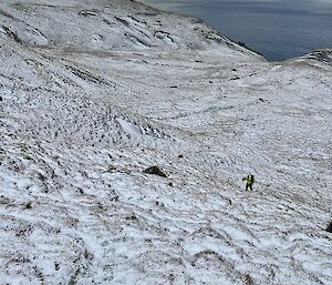 In the distance, a person carrying pack walking up a steep snow covered, grassy slope