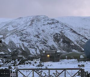 Front gate of station, reading "MACCA" in foreground, with station building and snow covered mountain in the background. Sunset light gives everything a pink glow