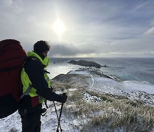 Man in field gear and wearing pack stands on top of hill looking back across the isthmus to station