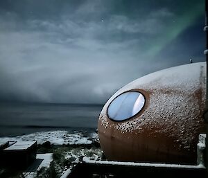 Brothers point hut, which is a large orange smarty shape, in the foreground, with sea in distance and small green aurora overhead