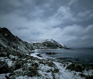 Mountains running into the sea, with snow covered track, very dark grey cloudy skies and sea tones