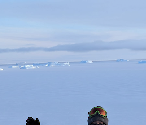 A man in yellow with a face muff on stands with arms outstretched on a rocky hill with snow in the background