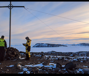 Three figures in yellow stand in twilight near a high pole with snow and rocky outcrops in the background
