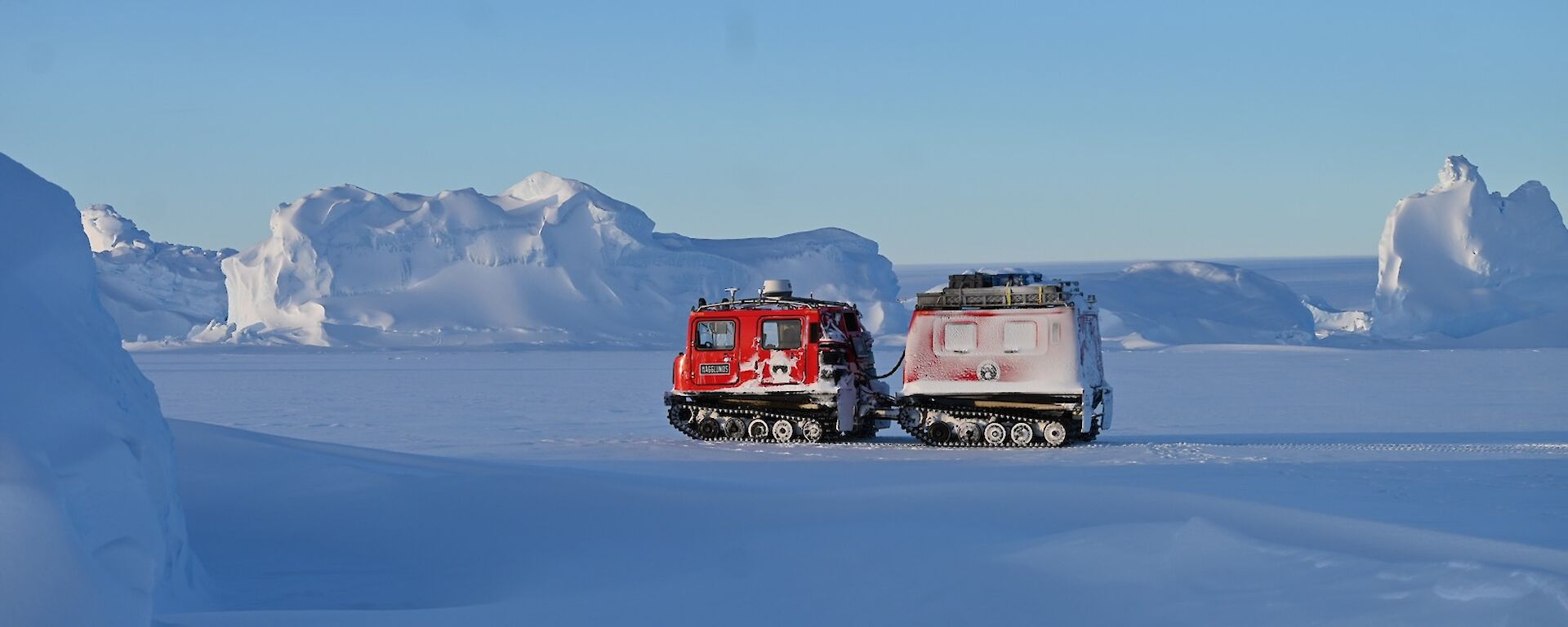 A red hagglund tractor stands on ice under blue sky with icebergs in the background