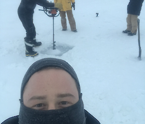 A man in the foreground with a face muff on and three fellows in the background drilling into the ice