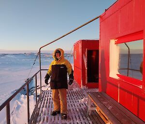 A man rugged up in yellow outdoor gear stands in front of a red hut