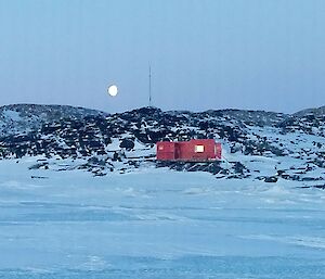 A red hut on a rocky hill with the moon in the background and snow in the foreground