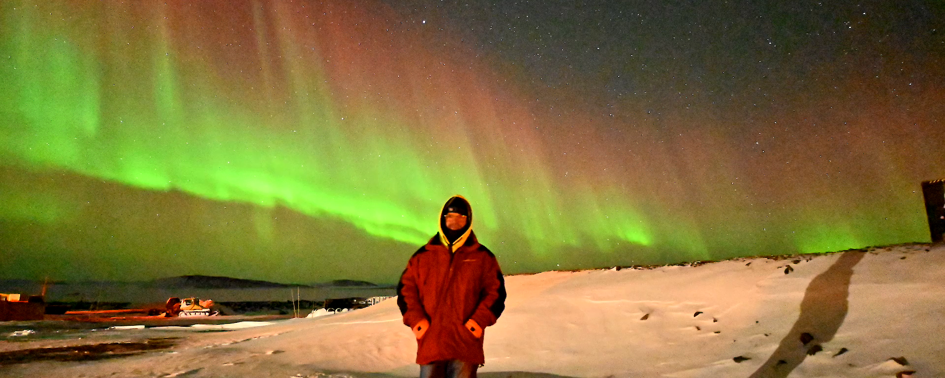 A man stands on snow with an aurora australis in the sky behind him