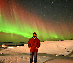 A man stands on snow with an aurora australis in the sky behind him