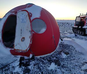 A red round hut with a window on a snowy slope with a red haggland behind it