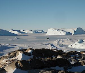 A green hagglunds on the sea ice driving towards Macey hut, with large icebergs in the background