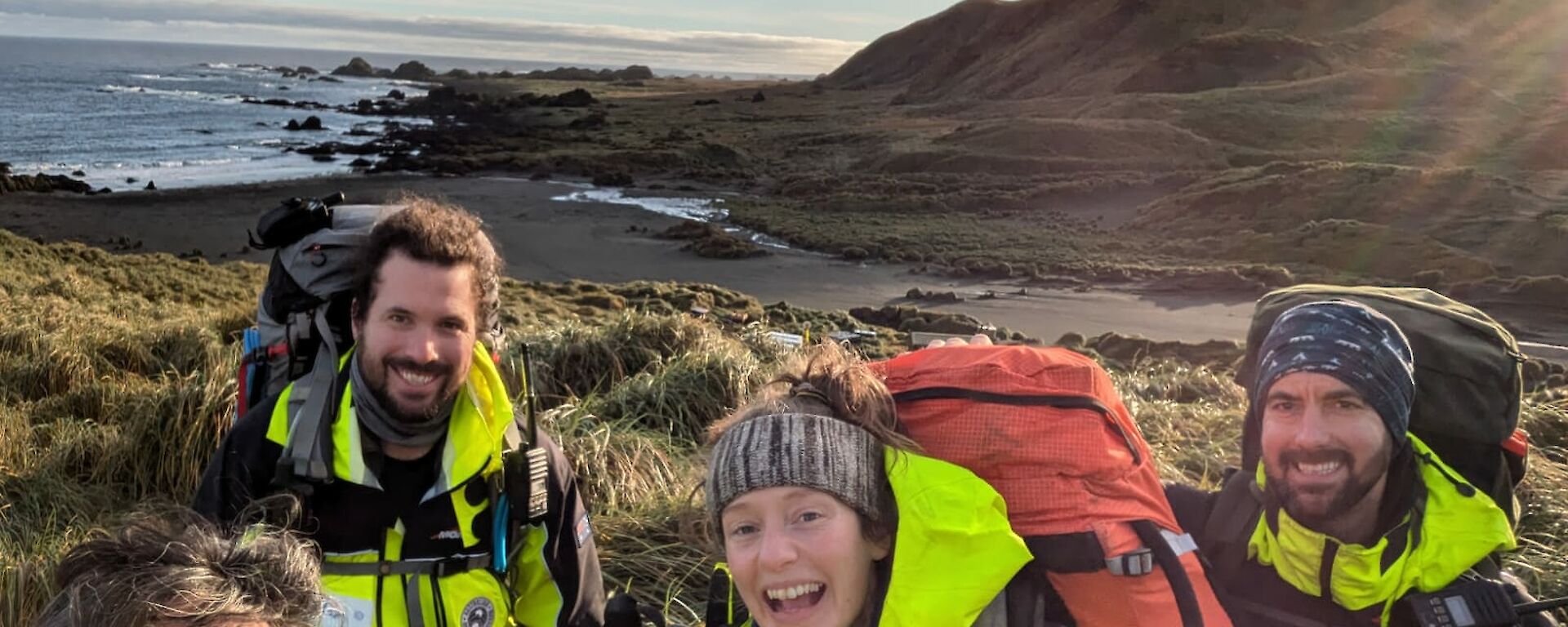 Four smiling people carrying packs with a green hill and sea behind them