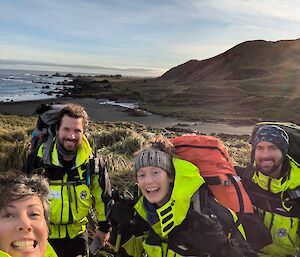 Four smiling people carrying packs with a green hill and sea behind them