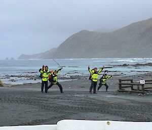 Four people with arms outstretched stand on a grey beach in front of a misty steep hill