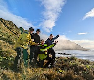 Four people stand on a green tussocky hill under blue sky