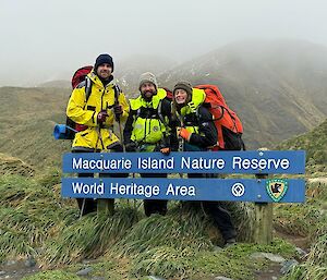Three people in yellow outdoor gear stand in front of a sign saying 'Macquarie Island Nature Reserve World Heritage Area'