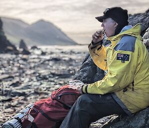 A person sits on a rock and looks out at a rocky beach and steep hills