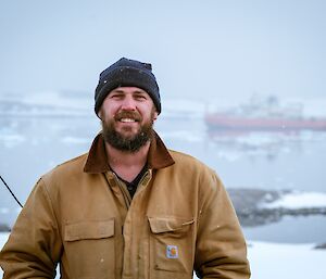 a smiling expeditioner stops for a portrait with the RSV Nuyina supply vessel shown through the backdrop of a foggy sky with calm glass like water and small icebergs