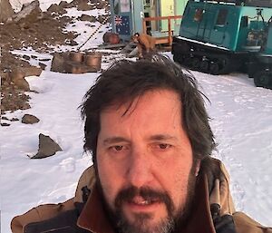 A man with dark hair and a beard takes a selfie in front of a green Hagg and shed