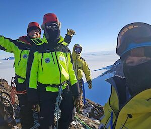 Three people standing on a cliff side with snow behind them, dressed in yellow Antarctic gear.