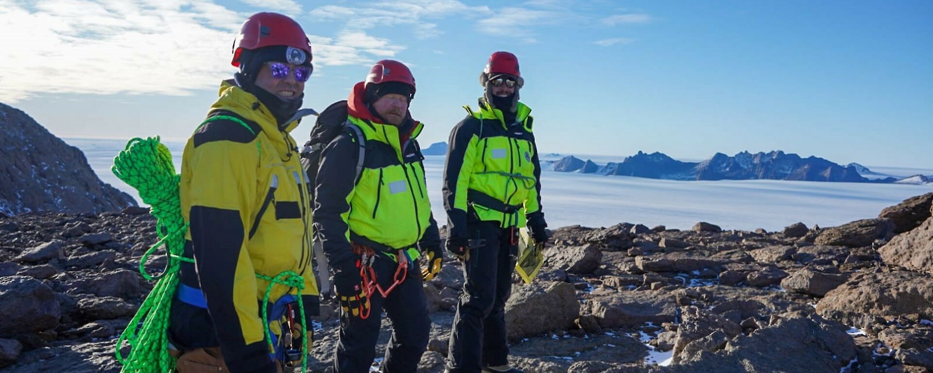 Three people in yellow on a rocky outcrop in a plain of snow under a blue sky