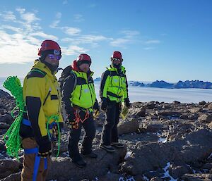 Three people in yellow on a rocky outcrop in a plain of snow under a blue sky