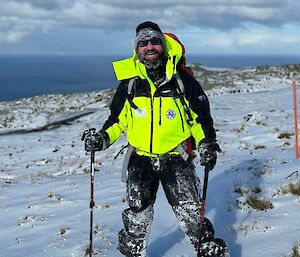 Icy news author, Cris, stands in snow wearing field kit and pack, holding walking poles, smiles to camera