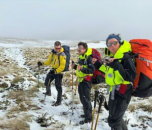 Three people wearing field kit, packs and sunglasses, stand on snow covered walking route amongst grass tussocks